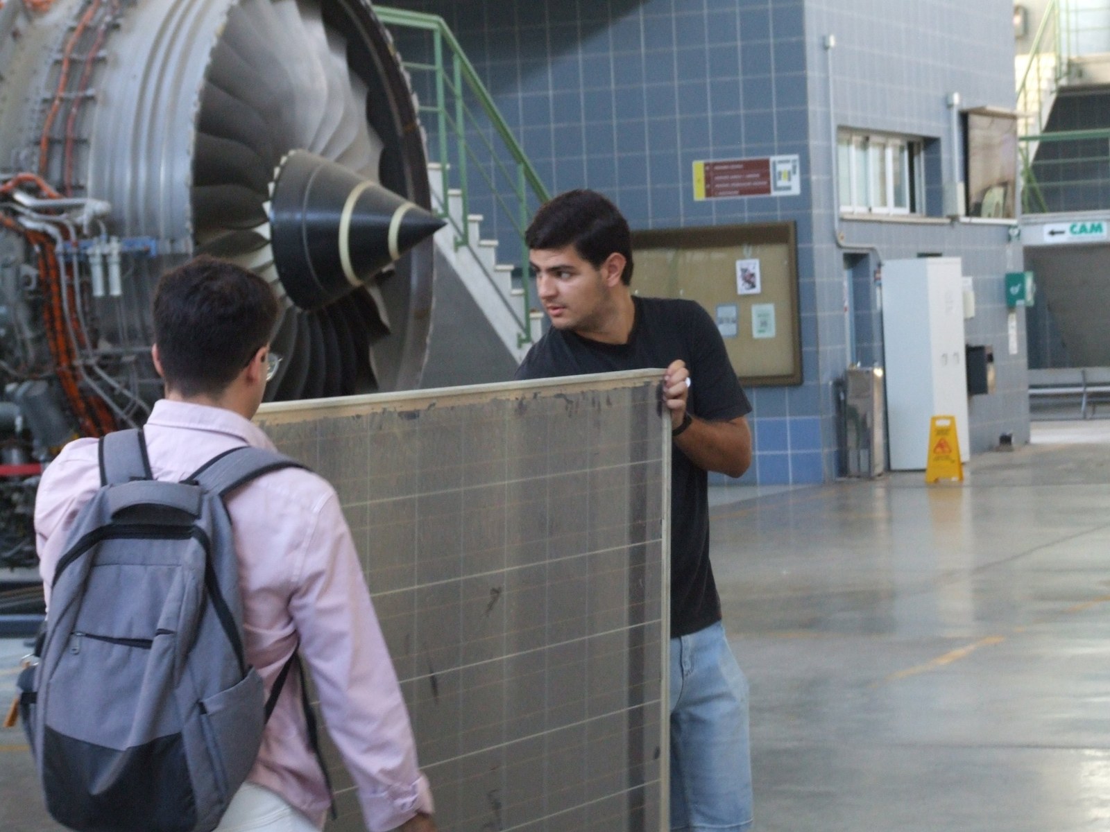 Dos estudiantes trasladando un panel fotovoltaico durante una visita a un hangar con un motor de avión